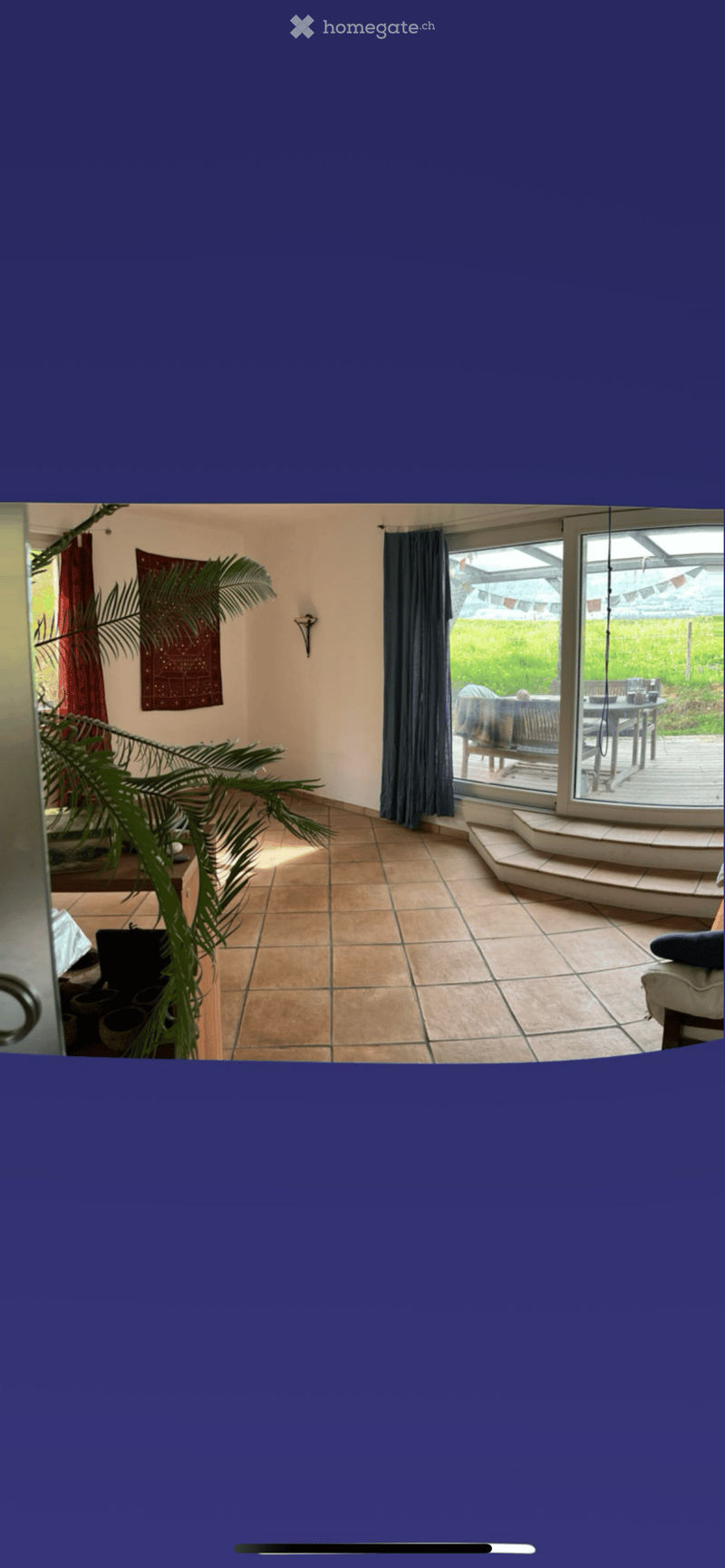 Bright living room with tiled floor, large sliding glass door leading to a patio, sofa, table, chairs, and a plant near the entrance.