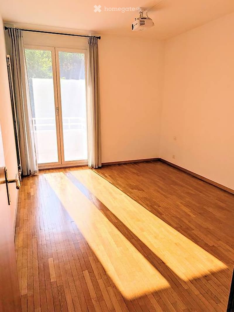 Empty room with wooden flooring, large sliding glass door with curtains, and natural light streaming in.