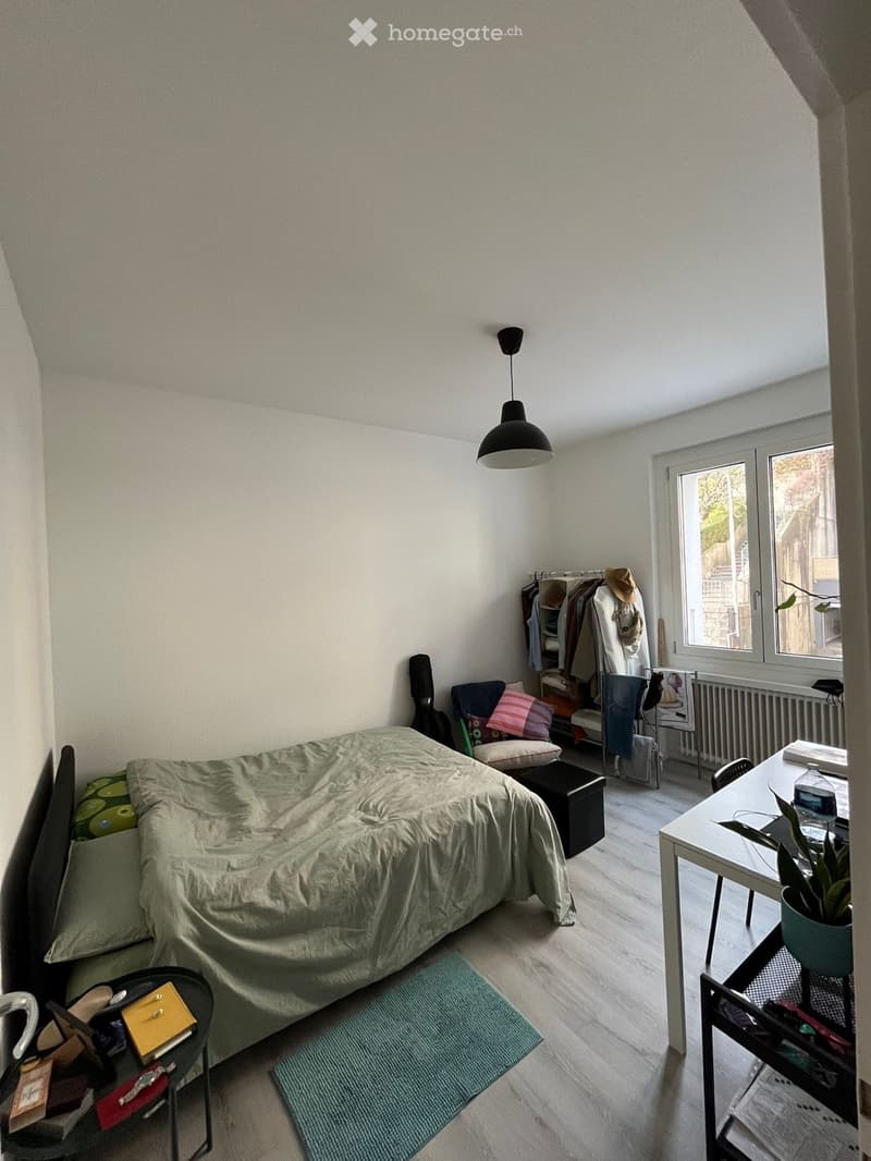 A small, well-lit bedroom with a bed, a desk, a chair, a stool, a plant, a radiator, a guitar, and a wardrobe.