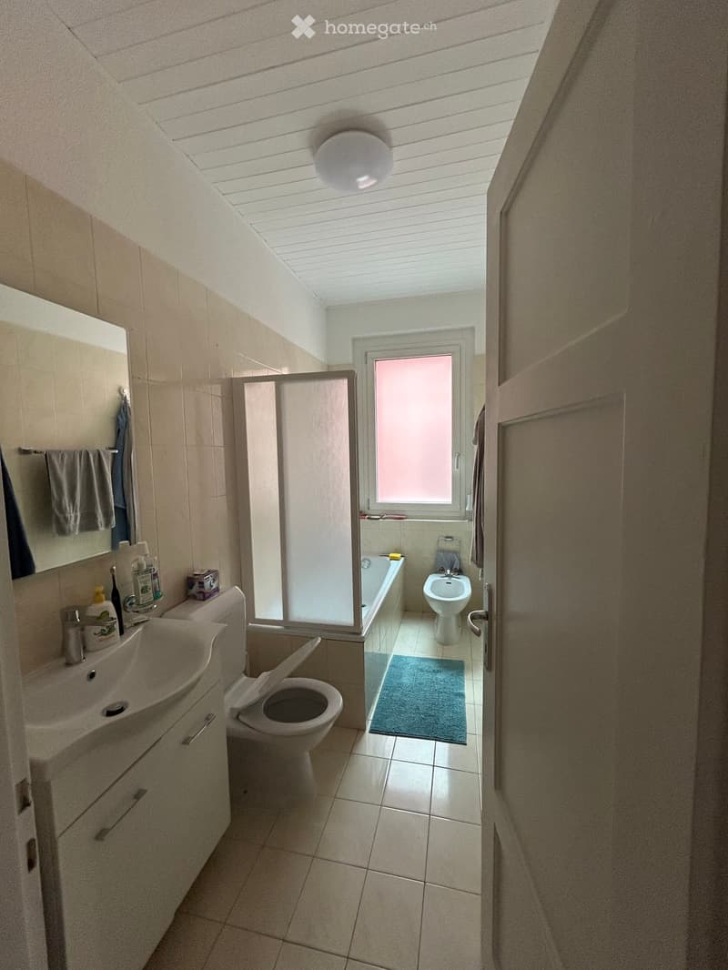 Bathroom with a sink, toilet, bathtub, mirror, and a shower stall. The room features tiled walls and floor, a window with frosted glass, and a small blue rug in the shower area.