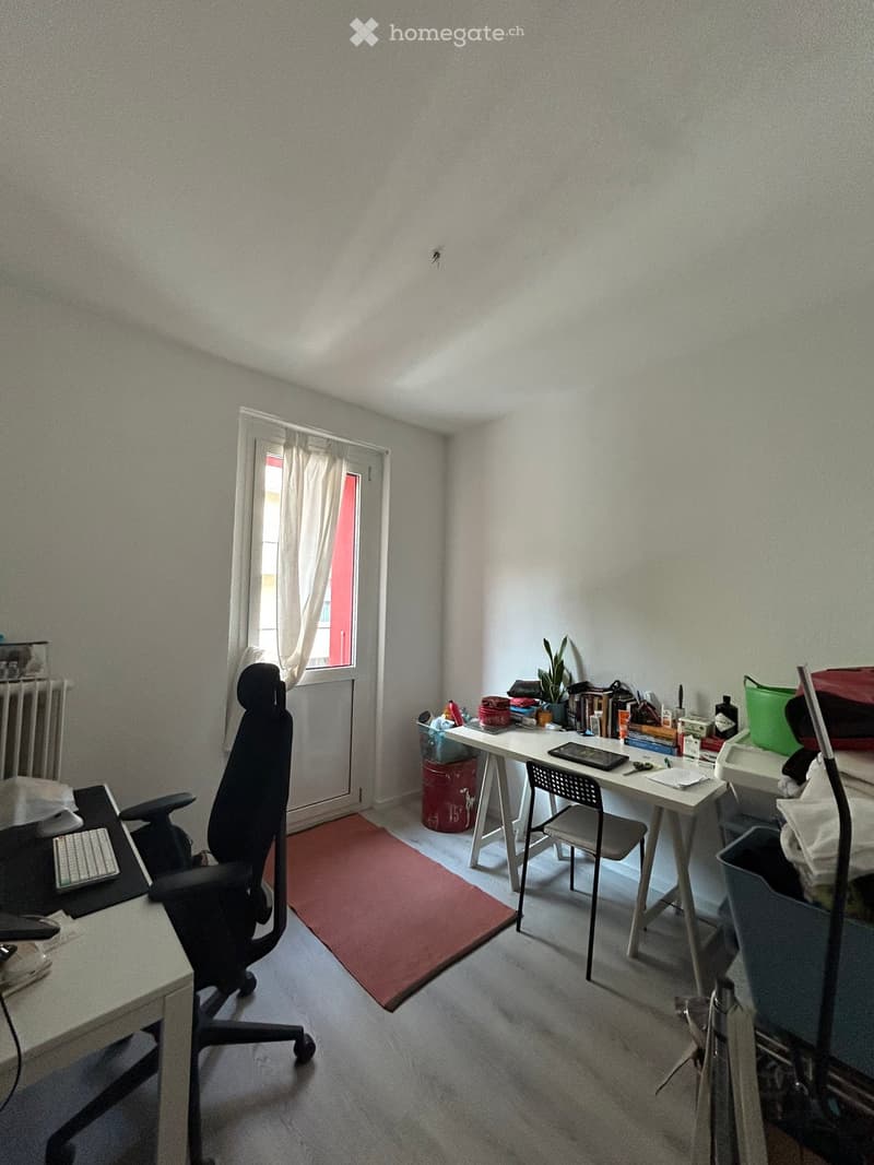 A furnished room with a desk, chair, laptop, keyboard, and a small plant on a side table. The room has a window with a curtain and a radiator below it.