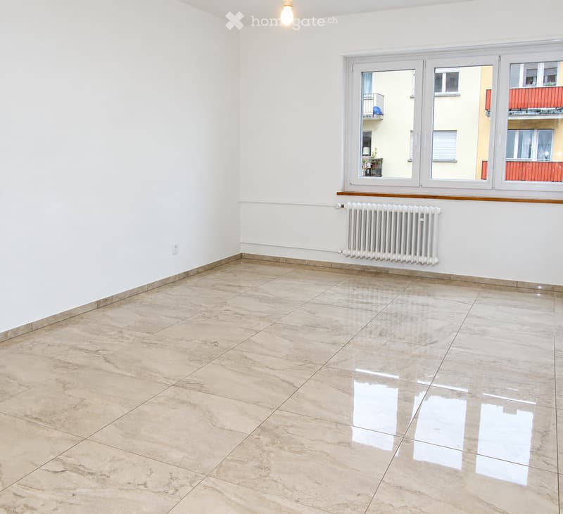 Empty room with white walls, tiled floor, radiator, and large windows showing a view of neighboring buildings and balconies.