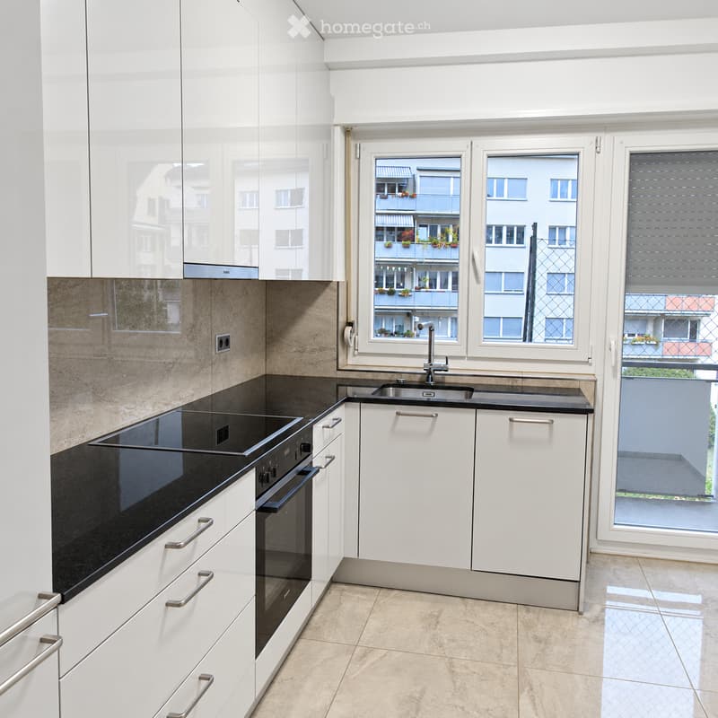 Modern kitchen with white cabinets, black countertops, stainless steel sink, and built-in oven. Large windows offer a view of the city.