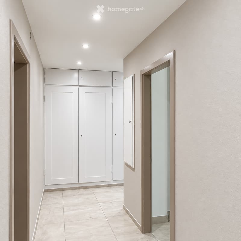 Clean, modern hallway with beige tiled flooring, white walls, and recessed ceiling lighting. Features built-in white storage units and a glass-paned door leading to another room.