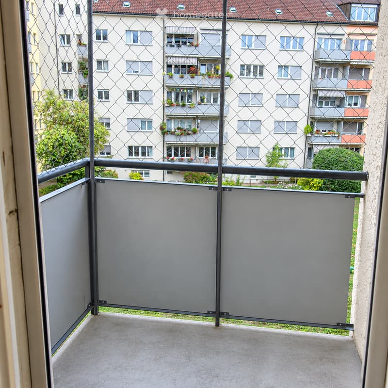 Balcony with grey privacy screens, metal railing, and a view of a multi-story residential building with balconies and greenery.