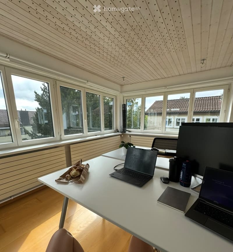 Bright, modern room with large windows, wooden floor, white table with laptops, screen, and office supplies.