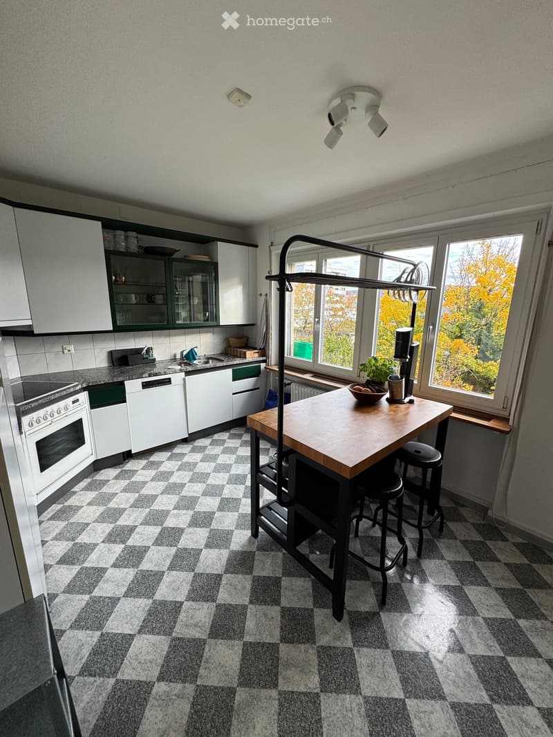 Modern kitchen with white cabinets, checkered floor, central island with stools, and large windows overlooking a leafy view.