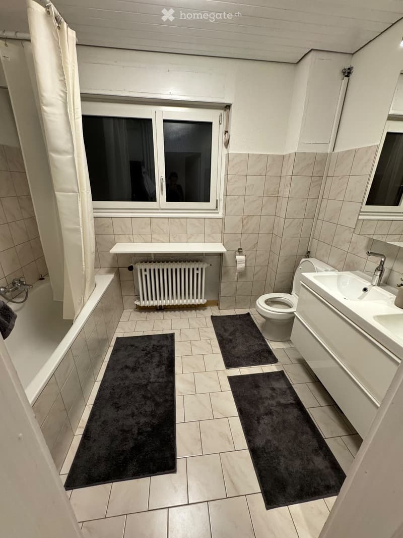 Modern bathroom with tiled walls and floor, featuring a bathtub, toilet, sink, radiator, and multiple black bathroom rugs.