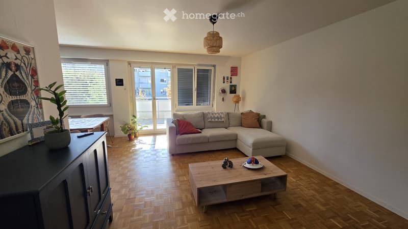 Bright living room with wooden flooring, large windows, and a sliding glass door leading to a balcony. Features a grey sectional sofa, wooden coffee table, side table, and potted plants.
