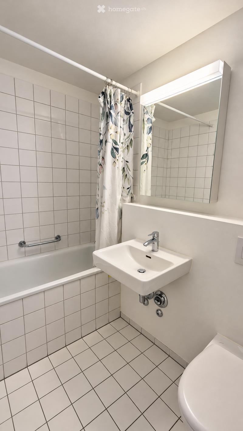 Modern bathroom with white tiled walls and floor, featuring a bathtub, sink, toilet, and a large mirror with built-in lighting. A floral-patterned shower curtain is visible.