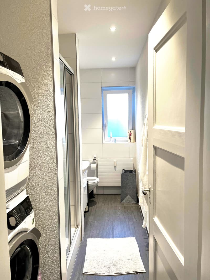 Modern bathroom with a washing machine, toilet, bathtub, sink, and a small window letting in natural light.
