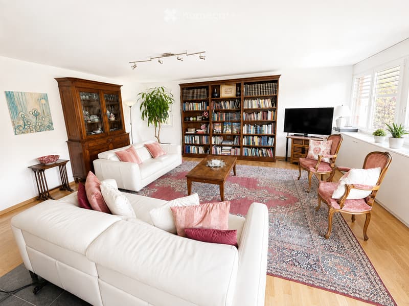 Bright living room with white walls, wooden floors, and large windows. Features a white sectional sofa with pink cushions, a wooden coffee table, two armchairs with pink cushions, a bookshelf filled with books, a TV, and a side table with a lamp.