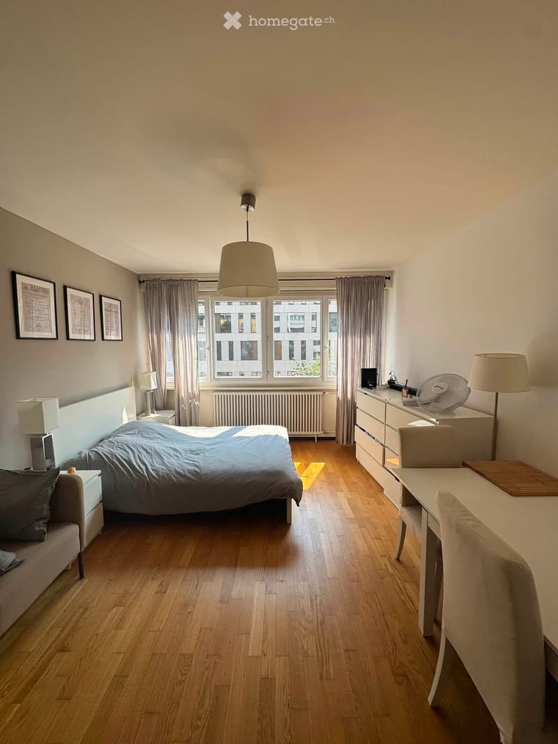 A well-lit bedroom with wooden flooring, a large bed with a blue duvet, a nightstand with a lamp, a desk with a chair, and a fan on top of a dresser. The room features large windows with curtains and several picture frames on the wall.