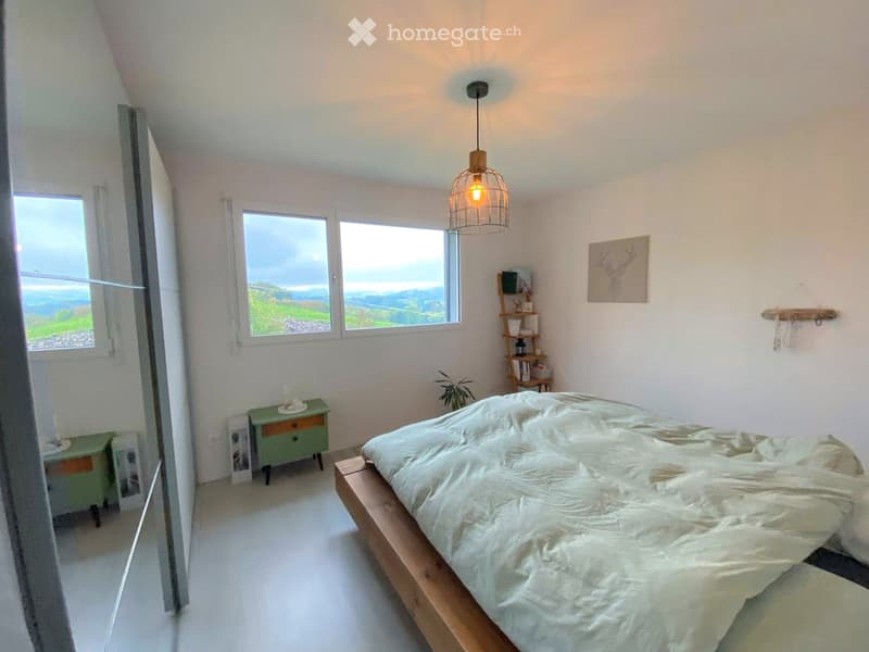 Bright bedroom with a wooden bed, nightstand, hanging lamp, shelf, and scenic view through large windows.