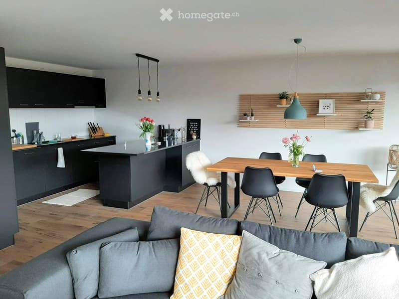 Modern open-plan living and dining area with a grey sofa, wooden dining table, black chairs, kitchen island, and pendant lights.