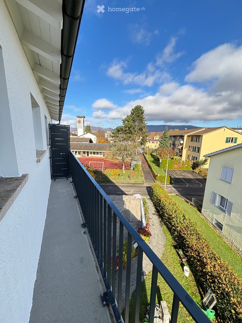 Balcony with metal railing, overlooking a residential area with houses, trees, and a distant view of mountains.