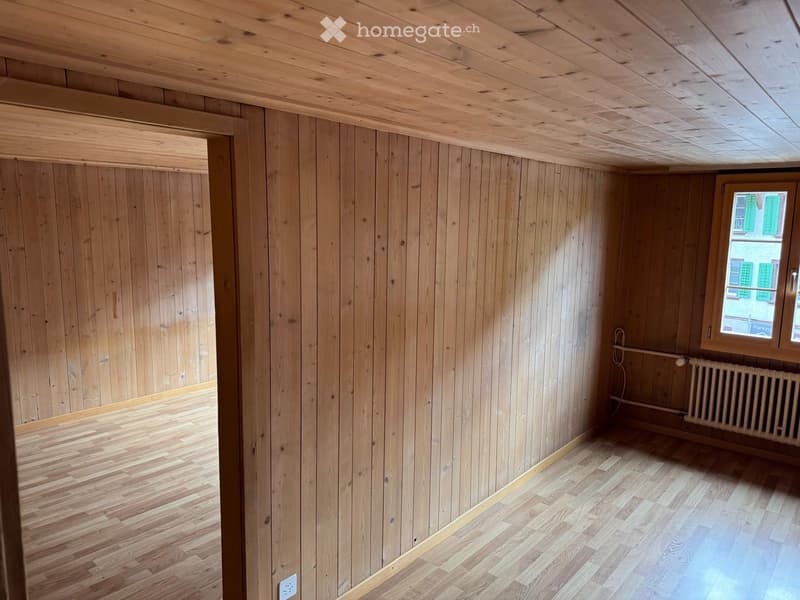 Empty room with wooden walls, wooden ceiling, and wooden floor. A window with a view of a building and a radiator are visible.
