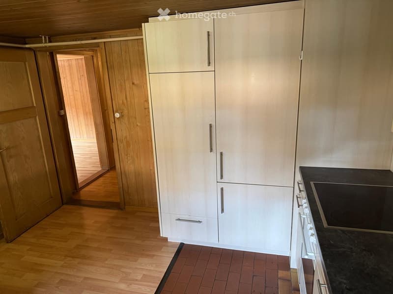 Modern kitchen with light-colored cabinets, a stainless steel refrigerator, and a black countertop with a built-in stove. Wooden flooring and a small tiled area near the cabinets.
