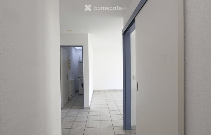 Empty hallway with tiled floor, white walls, and two doors leading to adjacent rooms. One door opens to a small utility area with a washing machine and dryer.