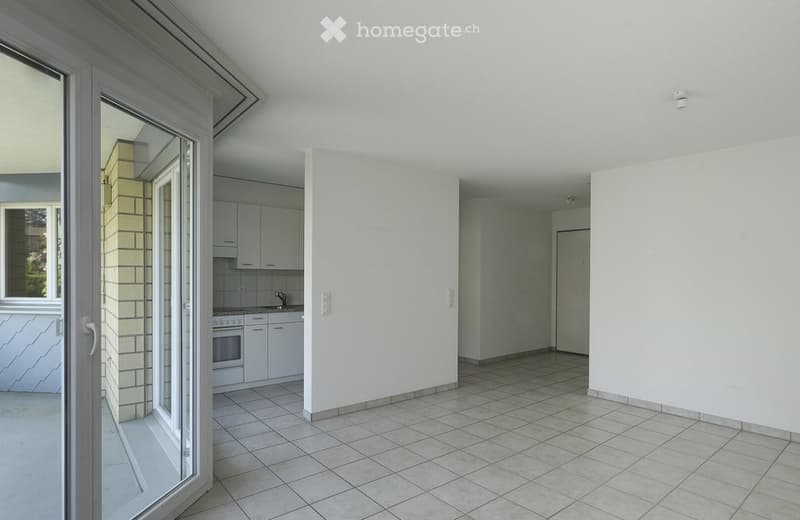 Empty room with tiled floor and white walls, connected to a small kitchen area with cabinets and appliances.