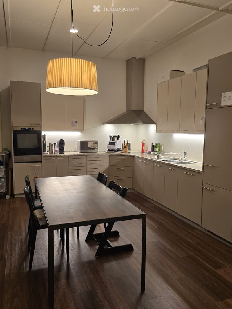Modern kitchen with light-colored cabinets, stainless steel appliances, a wooden dining table with chairs, and a hanging pendant light.