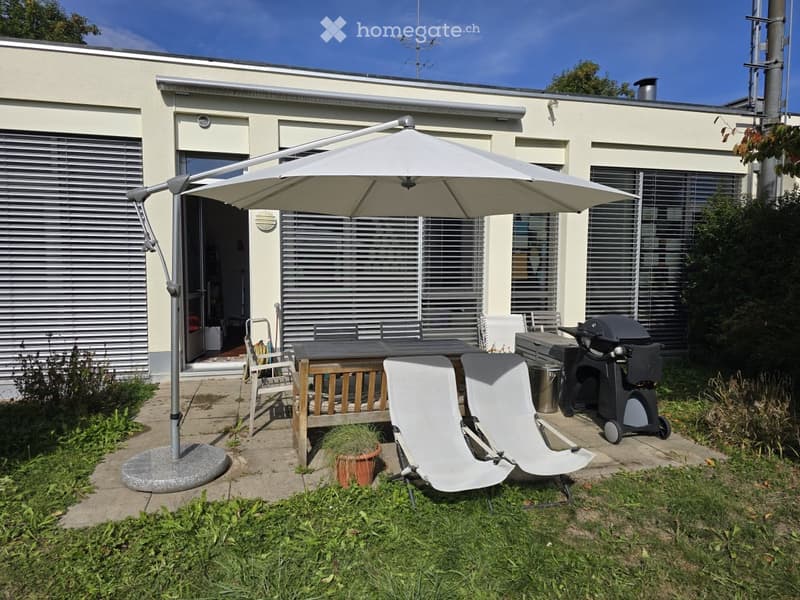 Outdoor patio area with a large umbrella, a wooden table, two white chairs, a grill, and a potted plant near the house entrance.