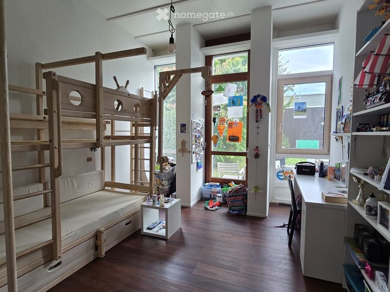 A well-lit room with a wooden bunk bed, a small sofa, a table, shelves, and a desk. The room has large windows and a wooden floor.