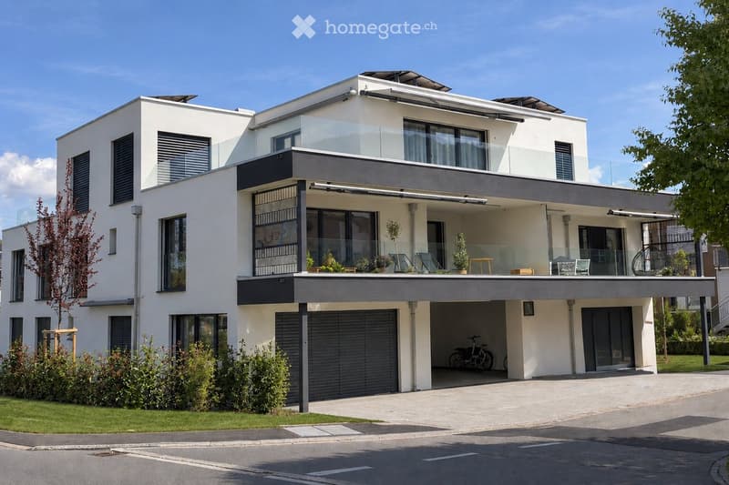 Modern two-story house with white facade, large windows, and a covered garage. Features a balcony with outdoor seating and plants, and a well-maintained garden area.