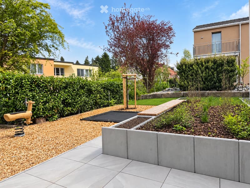 Well-maintained garden with a wooden seesaw, climbing frame, raised garden beds, and neatly trimmed hedges.