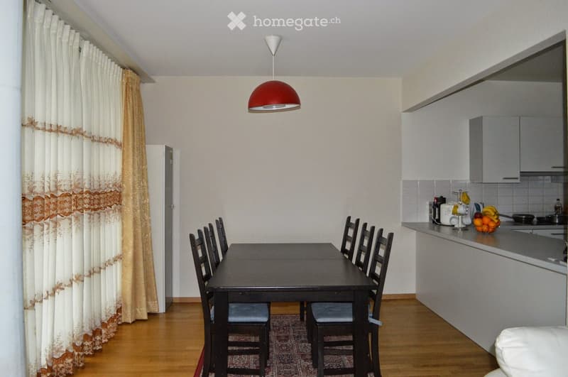 A well-lit room with a dining area and a small kitchen. The dining area features a black table surrounded by six black chairs with blue cushions. A red pendant light hangs above the table. The kitchen area has white cabinets, a countertop with a fruit bow