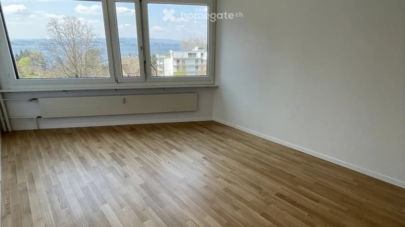 Empty room with light wooden flooring and large windows offering a scenic view of the water and surrounding area.