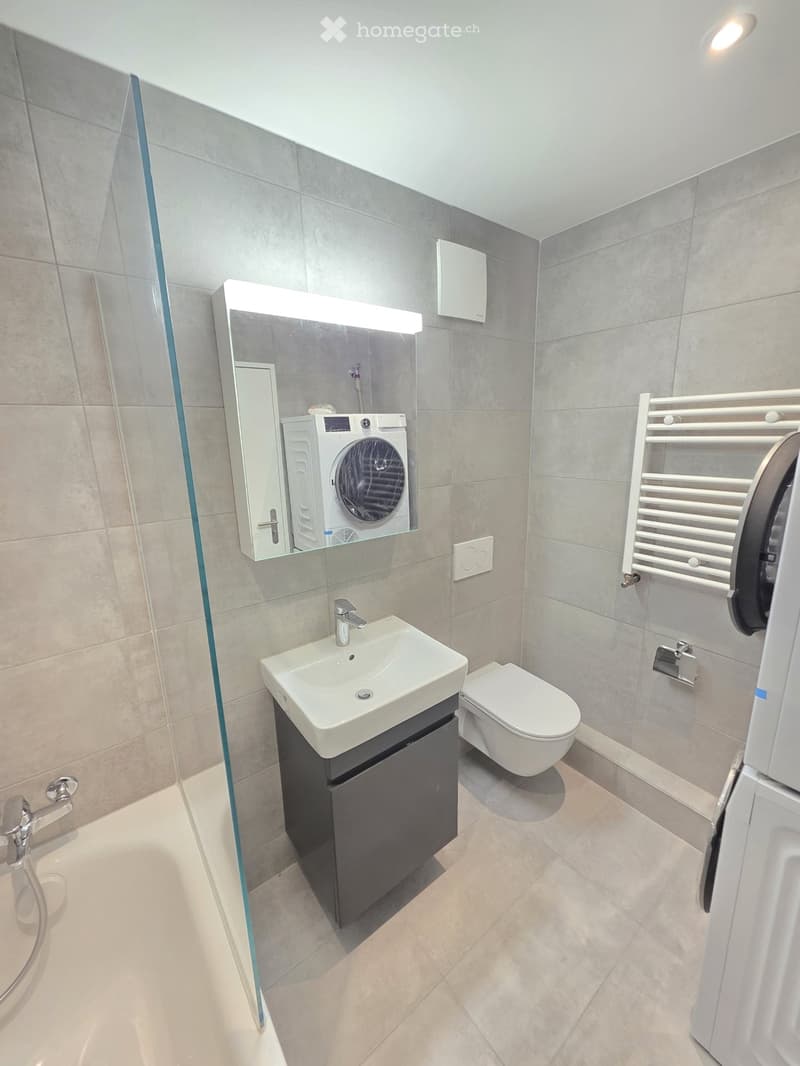 Modern bathroom with grey tiled walls, a white sink with a dark cabinet, a toilet, a shower with a glass door, a mirror with built-in lighting, and a radiator.