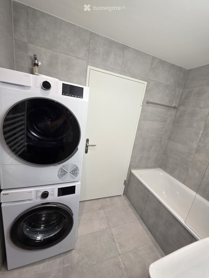 Modern bathroom with a white washing machine and dryer unit, a bathtub, and grey tiled walls and floor.