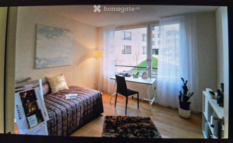 Cozy bedroom with a bed, desk, chair, lamp, and plant near the window. The room features a large window with a view of the city and a mirror on the wall.