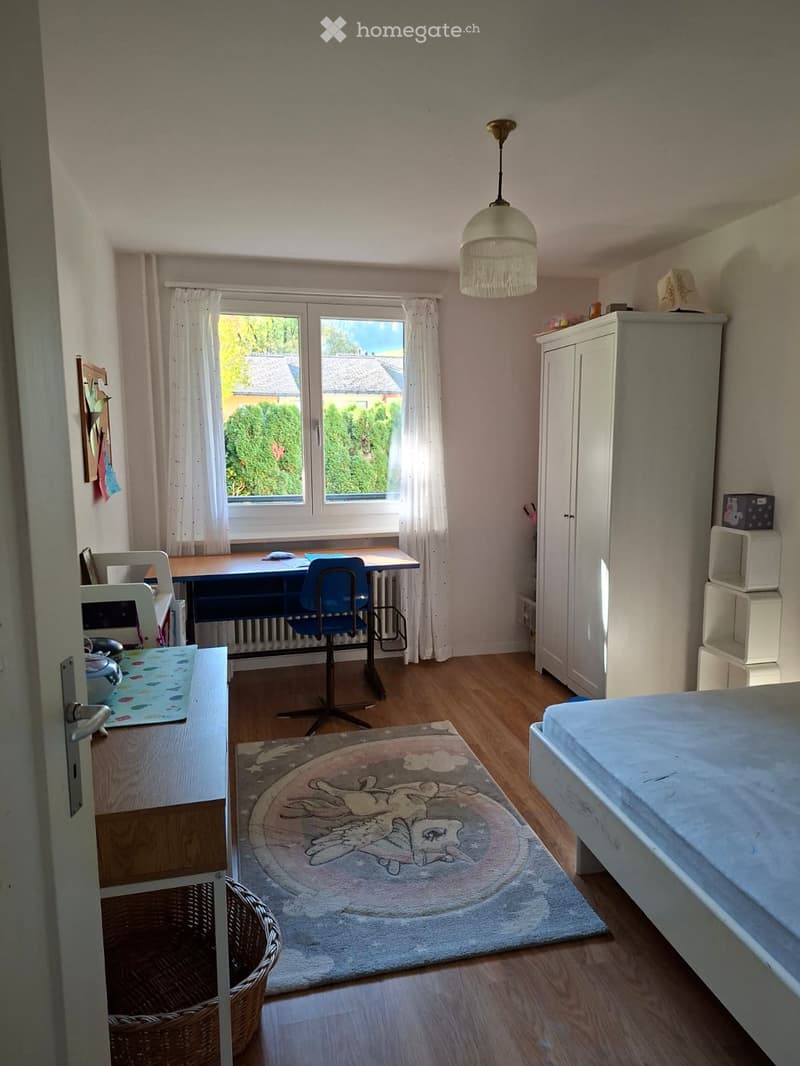 Well-lit bedroom with wooden flooring, a bed, desk, chair, wardrobe, and a round rug with a cartoon design.