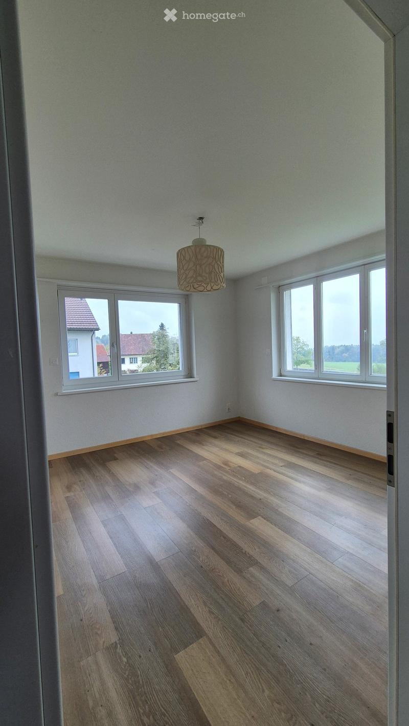 Empty room with wooden flooring, white walls, large windows, and a hanging light fixture.