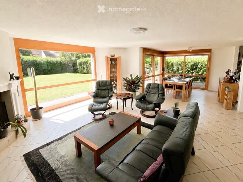 Bright living room with large windows, leather sofa, coffee table, armchairs, ottoman, dining area with table and chairs, and decorative plants.