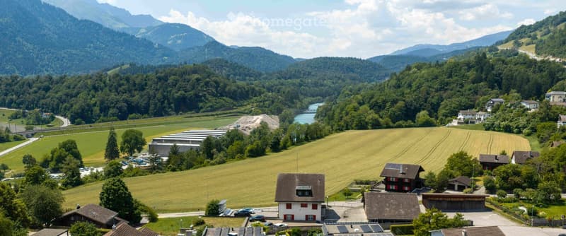 Aerial view of a rural village nestled in a valley surrounded by lush green fields and mountains. The village features traditional houses with red and white facades, surrounded by trees and open fields.