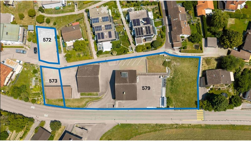 Aerial view of a residential area showing multiple buildings, parking lots, and surrounding greenery. Three plots are highlighted with blue outlines and labeled with numbers (572, 573, 579).