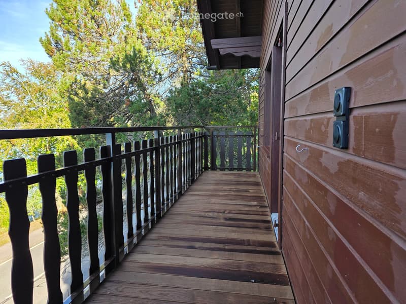 Wooden balcony with black railing, overlooking a green, tree-filled area.