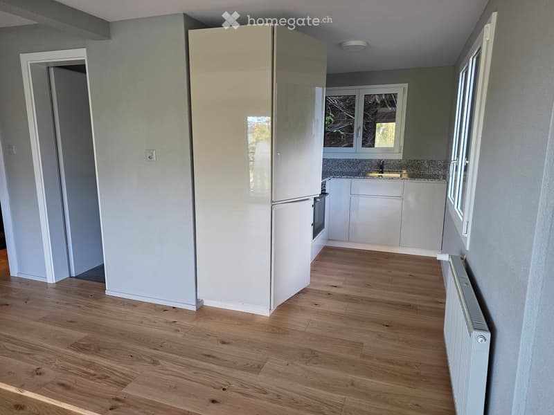 Empty room with wooden flooring, white walls, and a doorway leading to a kitchen area. The kitchen features white cabinets, a countertop, and a sink.