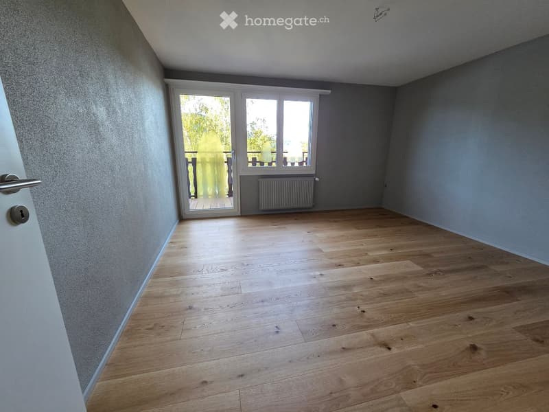 Empty room with wooden flooring, grey walls, and a large window with a view of a balcony area.