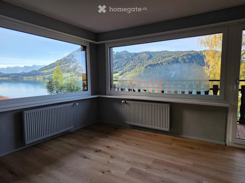 Empty room with wooden flooring, large windows offering a scenic view of a lake and mountains, and radiators along the walls.