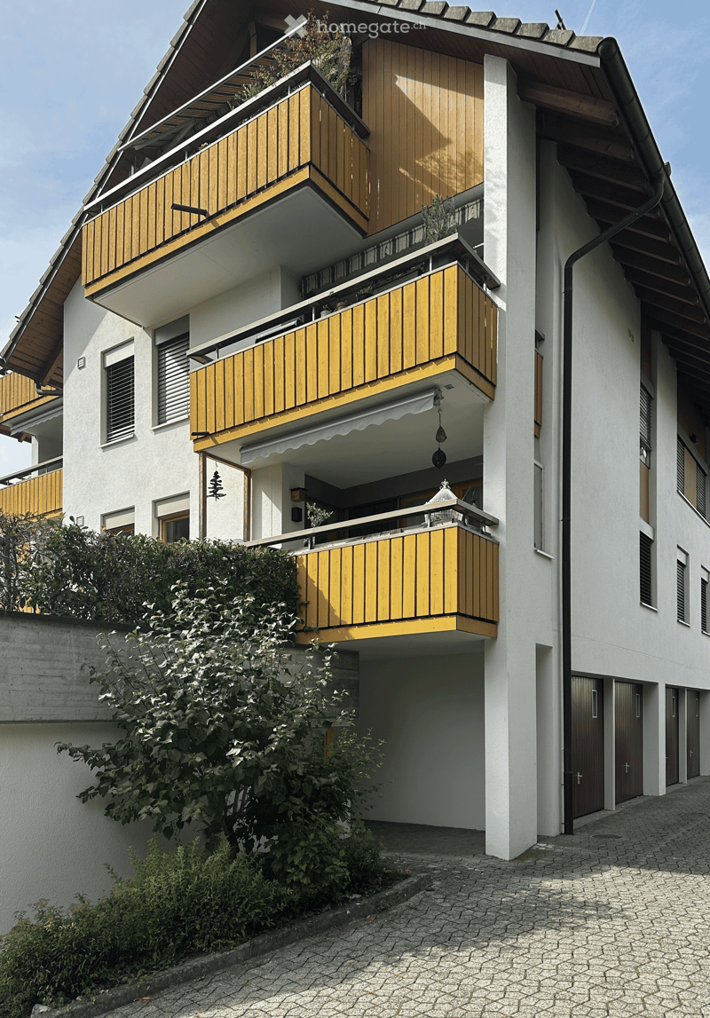 Multi-story residential building with white walls, wooden balconies, and multiple windows.