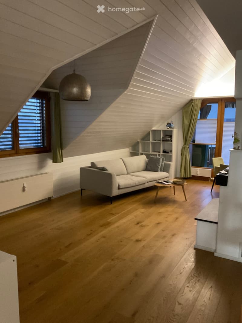 Bright living room with wooden flooring, a grey sofa, a small coffee table, a shelf with books, and a hanging pendant light.