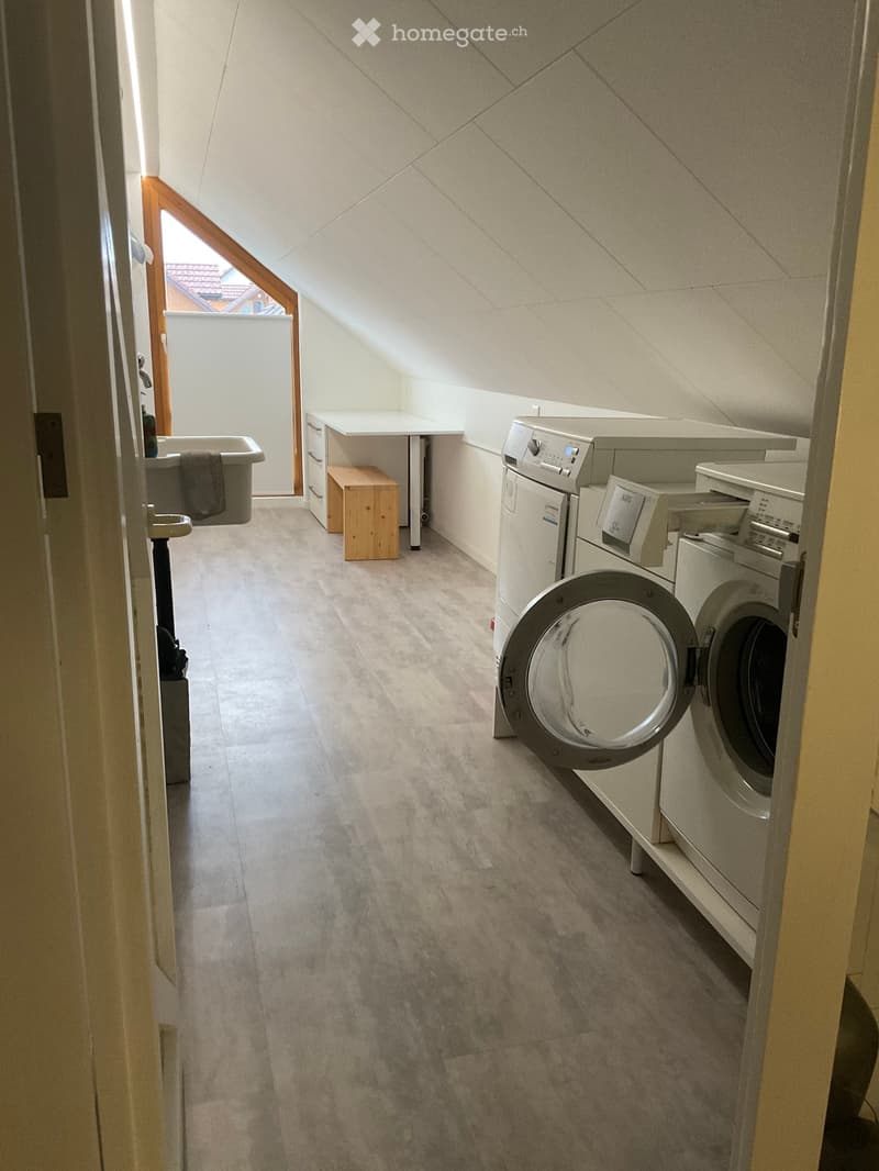 A small, well-lit bathroom with a washing machine, dishwasher, a small table, and a wooden stool. The room features a slanted ceiling and a window that allows natural light to enter.
