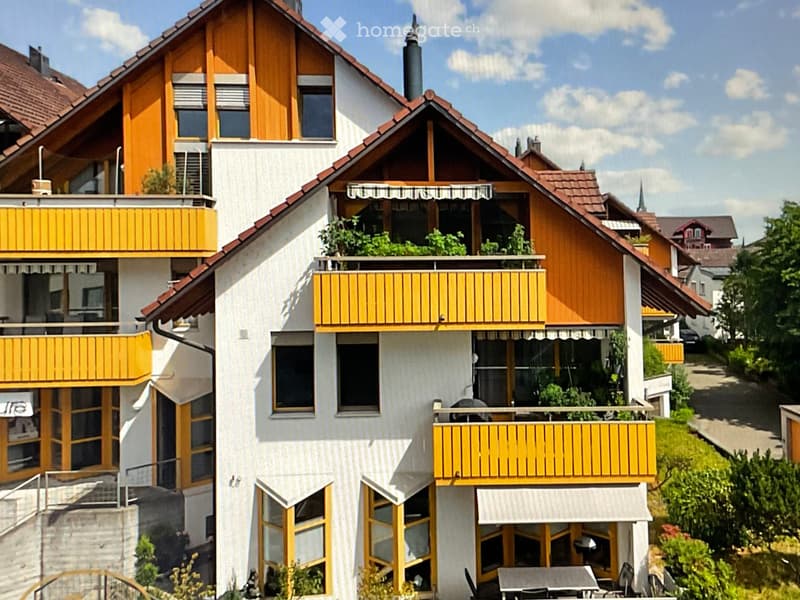 Multi-story house with white walls, yellow balconies, and wooden accents. Features multiple windows, a chimney, and a well-maintained garden area with outdoor furniture on the lower level.