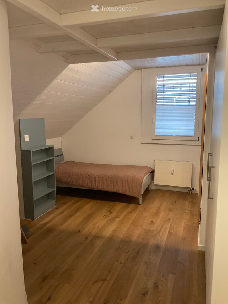 Cozy attic bedroom with wooden flooring, slanted ceiling, and a single bed with a pink duvet. A small shelf is positioned next to the bed, and a window with blinds allows natural light to enter.