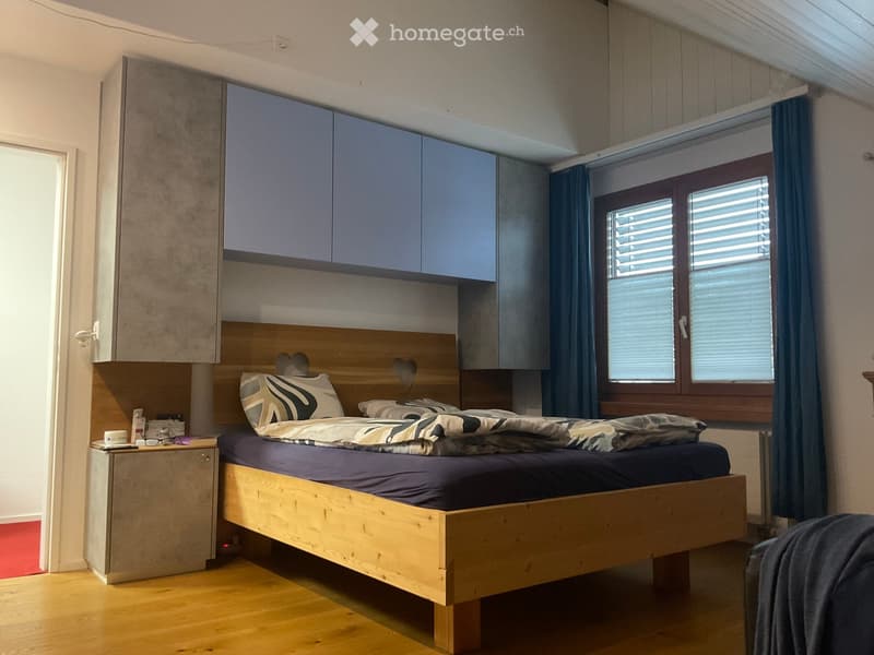 Modern bedroom with a wooden bed, nightstand, and built-in cabinets. Features a large window with blinds and a cozy bed with patterned bedding.