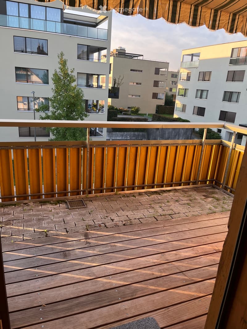 Balcony with wooden decking, metal railing, and brick-paved area, overlooking a modern residential complex.
